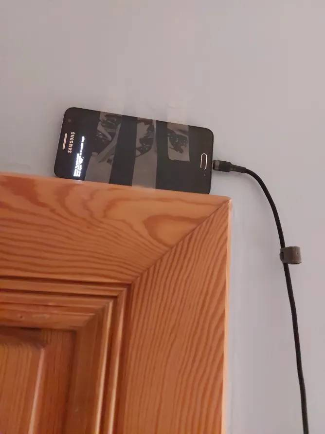 A picture of an old samsung phone duct-taped to a wall above a doorframe, with a charging cable going to it. On the display is the PostmarketOS console login prompt.