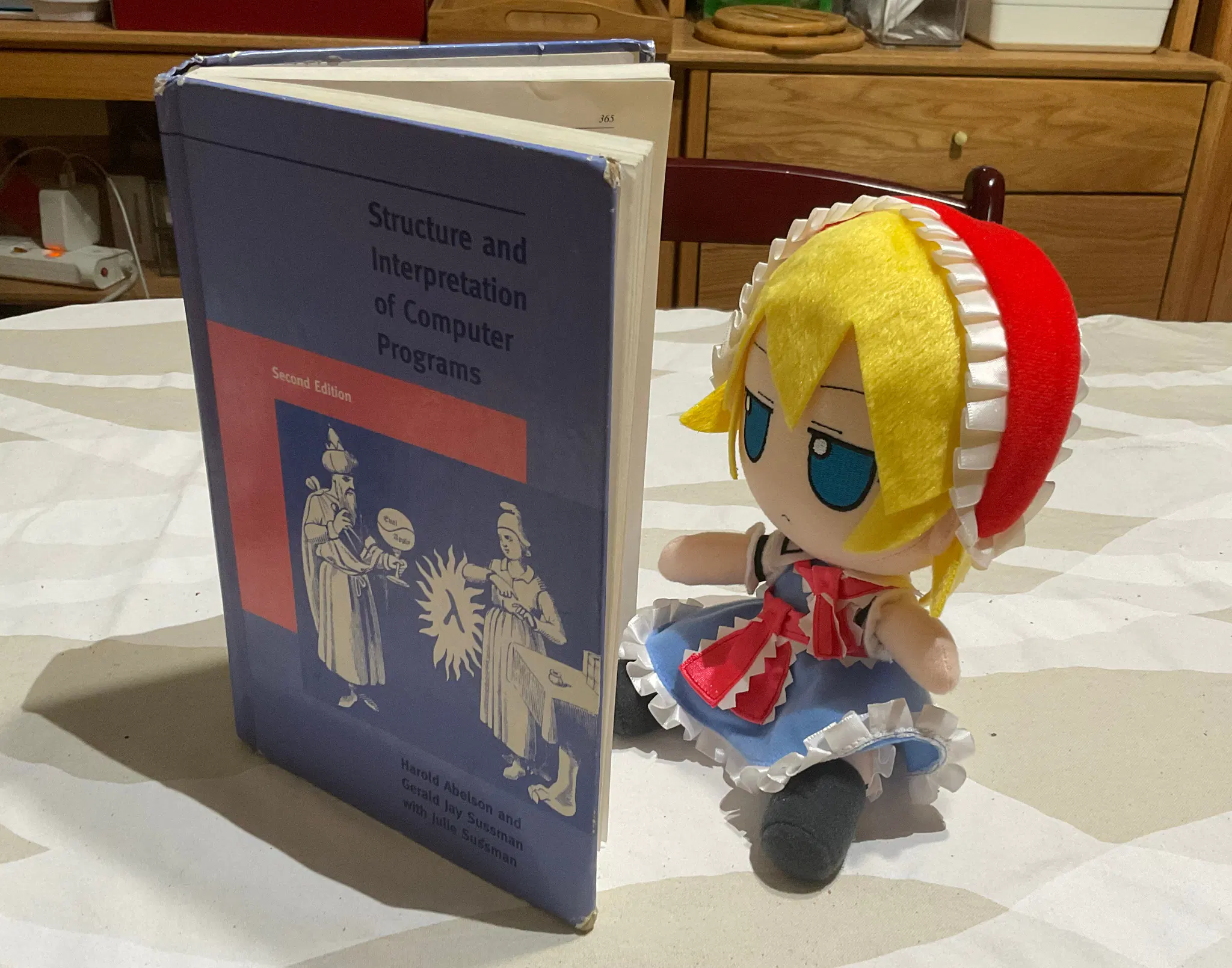 Fumo plush toy of Alice Margatroid from Touhou Project, behind an open book. The book "The Structure and Interpertation of Computer Programs" (SICP) with its iconic "wizardary" cover, as if Alice is reading it.