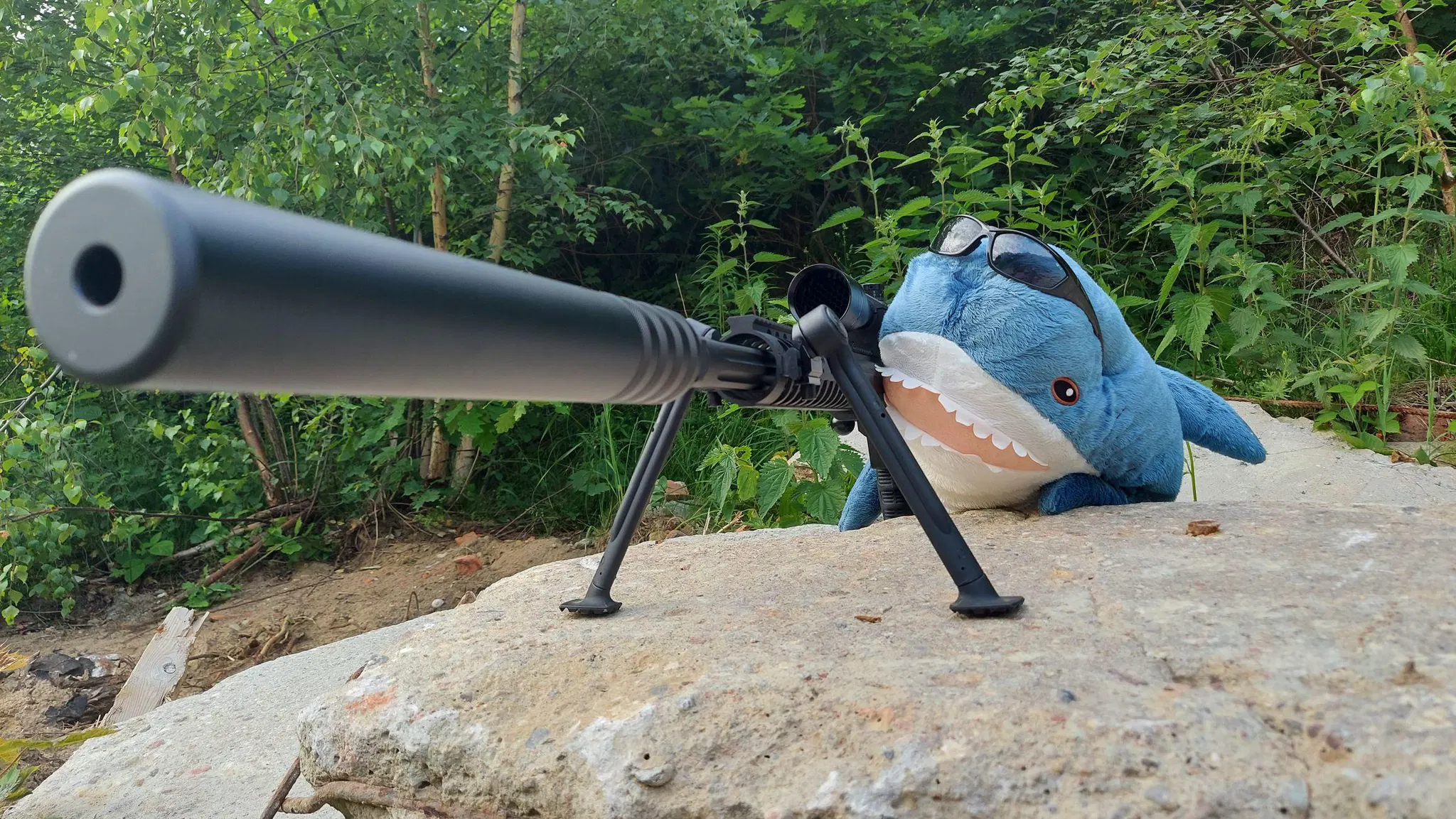Blåhaj plush wearing sunglasses on its head is humorously posed as a sniper, lying prone on a large, textured gray rock. The plushie is positioned behind a large, matte black sniper rifle equipped with a long suppressor and mounted on a bipod for stability. The entire scene is set outdoors, with a dense background of vibrant green trees and bushes, suggesting a forest or overgrown natural area